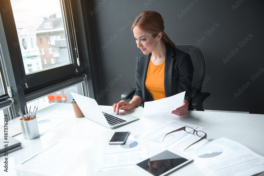 Poster side view smiling lady noting in keyboard of notebook computer and holding paper in hand. profession