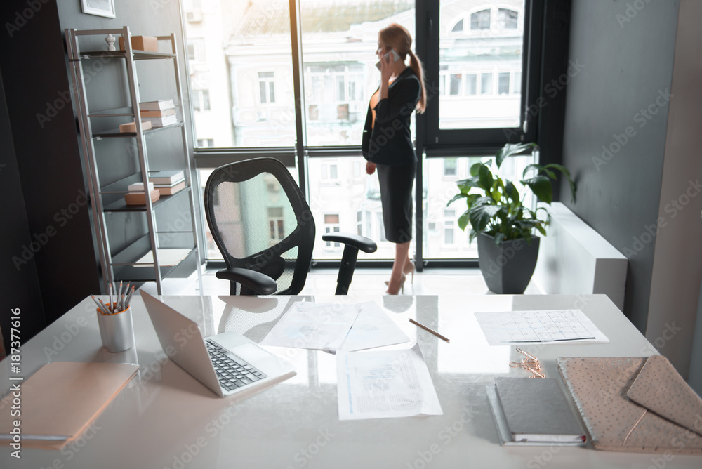 Poster full length side view serene businesswoman telling by mobile while situating near window in office. 