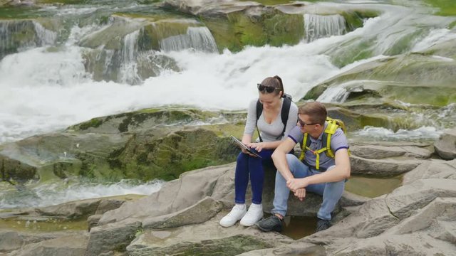 A young couple of tourists enjoy a tablet in a picturesque place near a mountain river. Technology and Tourism concept