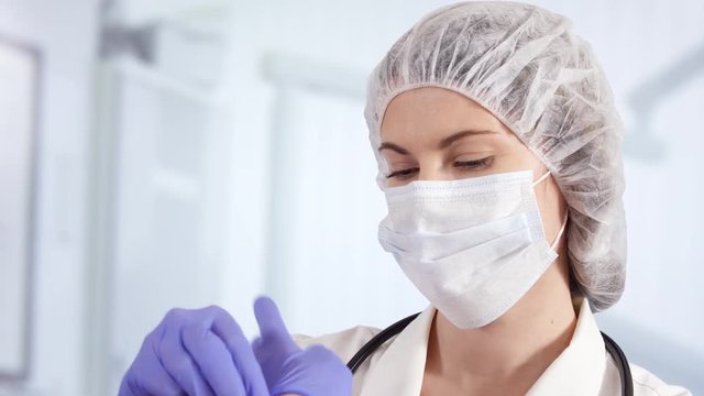 Confident professional female doctor in mask and cap in hospital room putting blue medical gloves on. Woman physician at work. Health care concept. Laboratory employee