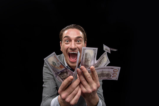 Passion about cash. Portrait of young insane man in suit is standing and showing a lot of money while looking at camera with desire and open mouth. Isolated on dark background