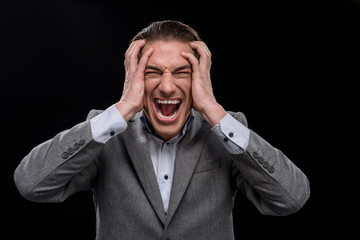 How is it possible. Portrait of angry young man is standing and touching his head. He is looking at camera while expressing annoyed and screaming. Isolated on dark background