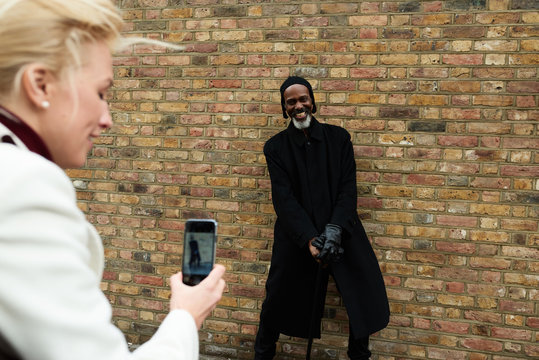Smiling man with cane posing against brick wall to woman with mo