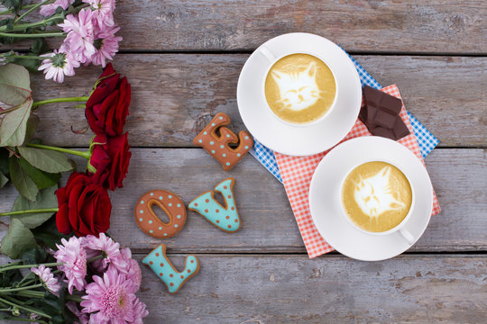 Flowers and cappuccino, old wooden background. Two cups of cappuccino with cats, word love from baked cookies and chocolate. Composition for Valentines Day.
