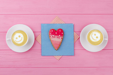 Red heart shaped cookie on napkin. Two cups of cappuccino with smile on pink wooden background, top view. Happy Valentines Day.