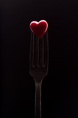 Red heart on fork, black background. Decorative heart and steel fork, still life. Valentines Day cutlery art.