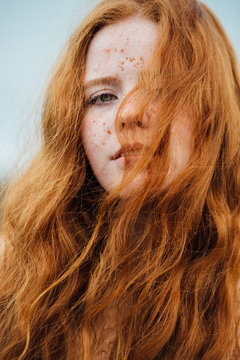Close up portrait of a young woman with ginger hair and freckles with her hair blowing over her face on a wintery day