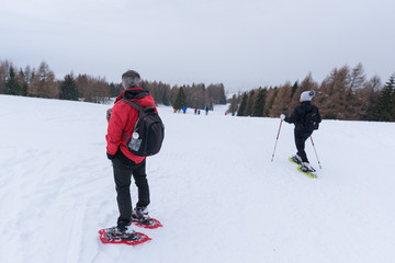 A mountain guide with graying hair looks at hikers as they walk on a snow-covered path while it snows. Man with red jacket, black backpack and rope. Generic, no face