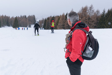 A mountain guide with graying hair looks at hikers as they walk on a snow-covered path while it snows. Man with red jacket, black backpack and rope. Generic, no face
