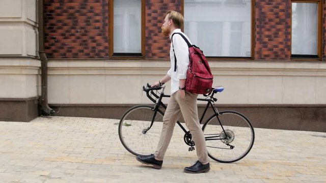 4k video of stylish hipster man with red beard walking on old street with vintage black bicycle