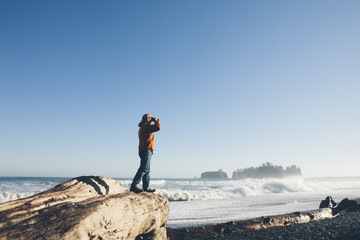 Man standing on log, looking through binoculars towards ocean & surf