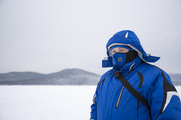 Fototapeta na wymiar Tourist in mask. A man in a blue winter jacket and mask on his face against the background of mountains in the snow. winter sports. Place for your text, design.