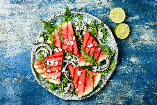 Fresh summer grilled watermelon salad with feta cheese, arugula, onions on blue background