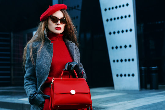 Outdoor portrait of young beautiful fashionable woman posing in street. Model wearing grey coat, red cashmere turtleneck, beret, cat eye sunglasses, holding handbag. Female fashion concept. Copy space