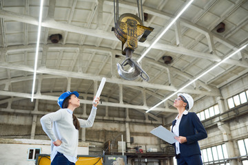Side view portrait two female engineers wearing hardhats examining workshop of modern factory looking at big crane hook, copy space