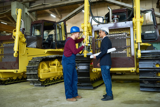 Profile view of young technician wearing overall and hardhat having informal working meeting with his superior while standing at spacious warehouse of modern heavy equipment factory.