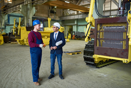 Smiling young technician wearing overall and hardhat sharing ideas with his superior while discussing promising project, interior of production department on background