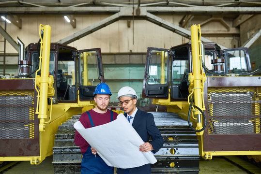 Handsome young businessman wearing classical suit and hardhat studying blueprint with help of industrial engineer while carrying out inspection at modern heavy equipment factory
