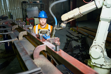 Head and shoulders portrait of bearded red-haired technician wearing reflective vest and hardhat operating machine, interior of production department on background