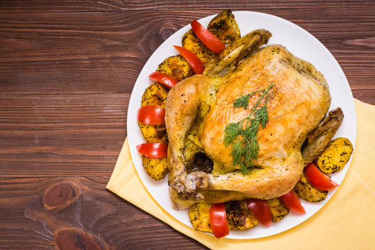 Baked whole chicken with a garnish of potatoes and tomatoes on a plate. Wooden table. Top view