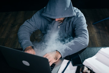guy hacker in a sweatshirt with a hood smokes and sits behind a laptop