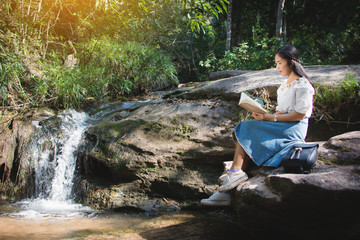 Asian women reading a book sitting on the rock near waterfall in forest background  , Relax time and hobby on holiday , soft focus,tone of hipster style