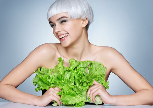 Smiling girl covering her breast a lettuce leaves. Photo of fashion blonde girl on blue background. Detox concept