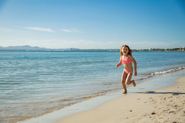 Fototapeta premium Cute happy little girl running along the beach in swimming suit, jumping over waves. Beautiful summer sunny day, blue sea, turquoise water, picturesque landscape. Majorca, Spain