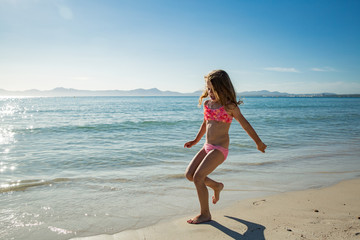 Fototapeta na wymiar Cute happy little girl running along the beach in swimming suit, jumping over waves. Beautiful summer sunny day, blue sea, turquoise water, picturesque landscape. Majorca, Spain
