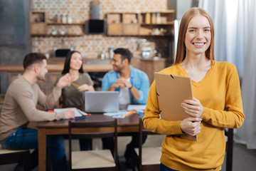 So pretty. Young beautiful positive woman standing with a folder in her hands while her calm pleasant skilled coworkers sitting at the table and talking