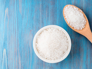 White bowl with large sea salt and wooden spoon on blue bright   wooden table. Top view.
