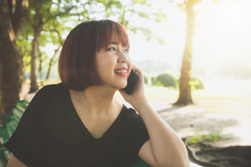 Cute asian woman smiles and talking on mobile phone while sitting in park spring day. Asian woman using on smart phone with feeling relax and smiley face. Lifestyle and technology concepts.