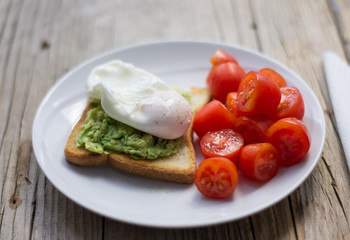 Perfect poached egg on a toast with smashed avocado and fresh tomatoes on a side on  a white plate on the wooden table