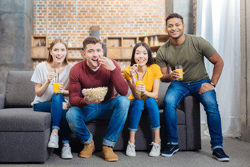 Amazing film. Positive friendly young people eating tasty popcorn and drinking orange juice while sitting on a sofa and watching an interesting film