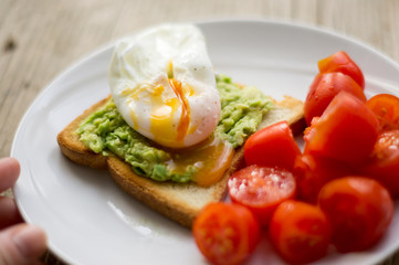 Perfect poached egg on a toast with smashed avocado and fresh tomatoes on a side on  a white plate on the wooden table