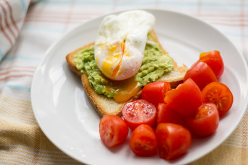 Perfect poached egg with avocado toast and fresh tomatoes on a white plate on a dish cloth