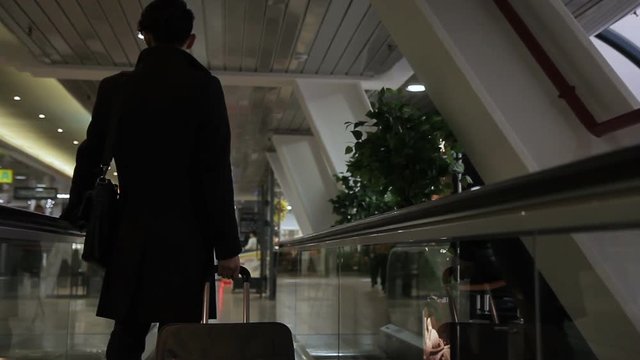 The asian businessman is standing on the speedwalk in the airport. The investor is wearing black coat and has a luggage and a bag while traveling. The man departs to the main hall to finish his flight