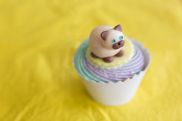 sweets, treatment, confection concept. on the yellow background of tablecloth there is marvelous cake in paper cup decorated with figure of cat that is lying on the top of whipped cream