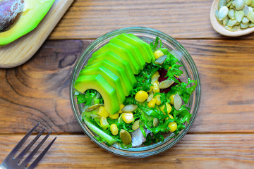 Vegetarian avocado lettuce salad in a glass bowl and on a wooden table. Salad with fresh avocado, lettuce leaves, canned corn and pumpkin seeds. Easy and useful recipe. Top view