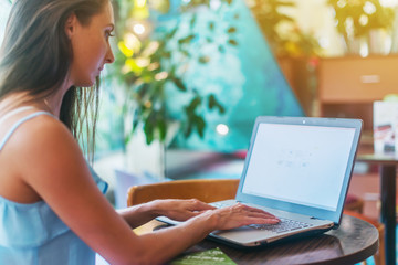 Young business woman working on her laptop, typing, sitting in a restaurant next to window sunny summer morning. Female writer using the netbook