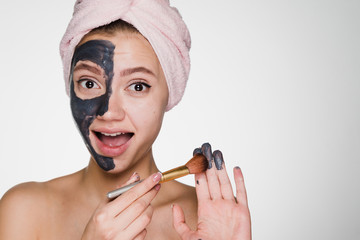 happy young girl with a pink towel on her head puts a clay mask on half of her face