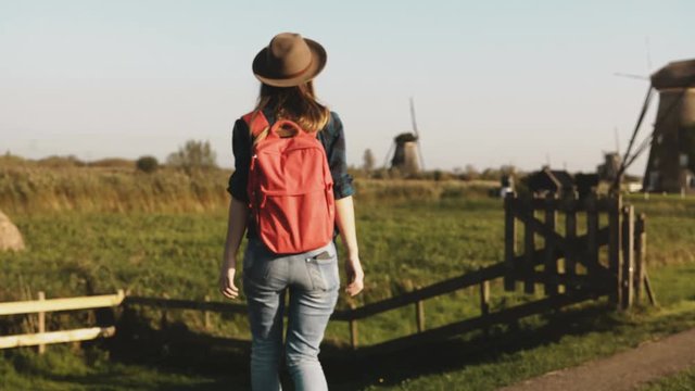 Casual girl jumps over fence near windmill farm. Woman in hat with long hair and red backpack returns home on sunset. 4K