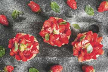 Summer and spring dessert. Home pies tartlets with custard and strawberries, decorated with mint and powdered sugar. On black stone table, rustic, with wooden board, tray. Copy space top view