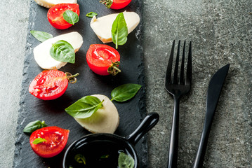 Italian dinner. Homemade salad caprese - tomatoes, fresh basil, mozzarella cheese, olive oil. On a black stone table. Served on skewers. Copy space top view