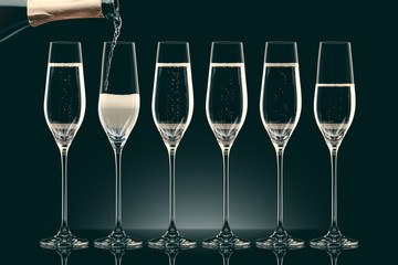 pouring champagne from bottle into six transparent glasses on black
