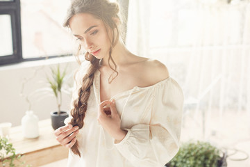 Sunny portrait of beautiful sexy and young woman with dark braid  wearing simple light dress in studio with big windows, plants, bath and straw chair