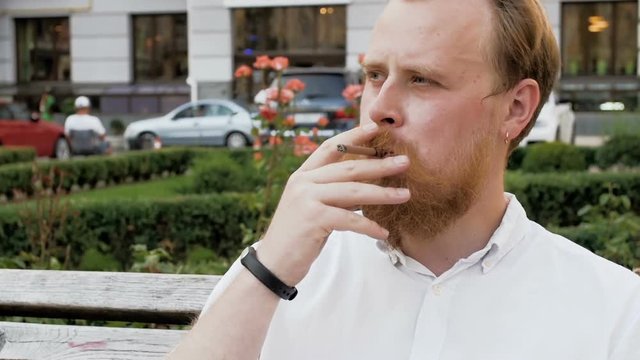 Closeup slow motion portrait of stylish red bearded man smoking cigarette at park