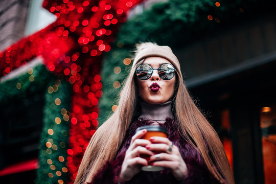 Outdoor portrait of young beautiful happy smiling hipster girl posing on street, looking at camera Model wearing stylish winter hat, holds in hands coffee dressed in sunglasses, having fun