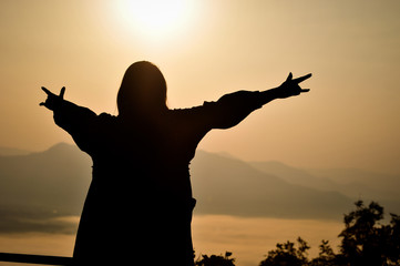 Silhouette of woman put her hand with 
Love symbol up in the air with background of early morning foggy sunrise on top of mountain soft focus.