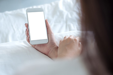 Mockup image of a woman's hand holding white mobile phone with blank desktop screen sitting in the white bed